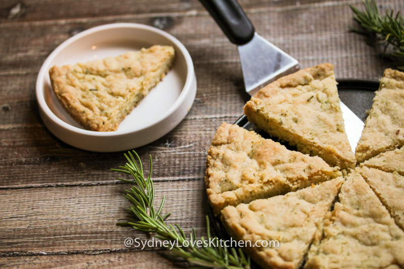 rosemary shortbread slice on a white plate and the whole pie of slices next to it and a couple of sprigs of rosemary on the side.