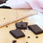 Easy Bake Oven Brownie Recipe with 3 brownies, a wooden spoon and some flour on the board, with a plate of brownies and a pink towel in the background