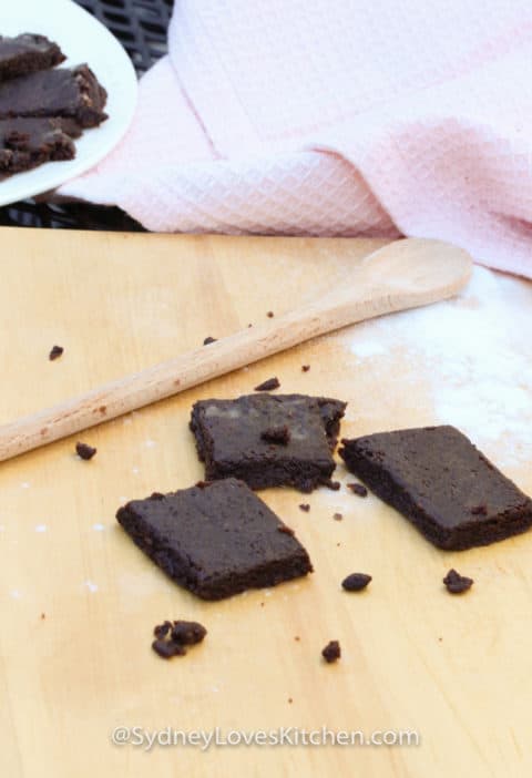 Easy Bake Oven Brownie Recipe with 3 brownies, a wooden spoon and some flour on the board, with a plate of brownies and a pink towel in the background
