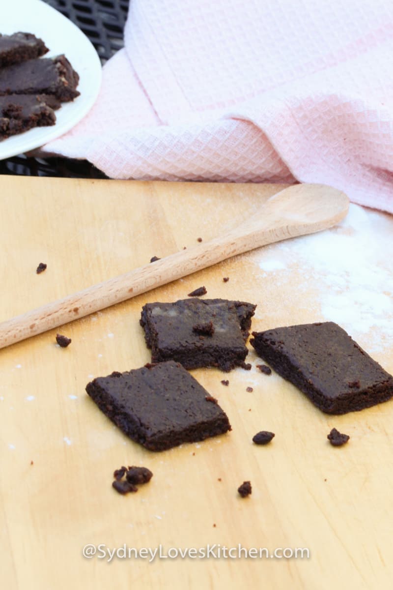 Easy Bake Oven Brownie Recipe with 3 brownies, a wooden spoon and some flour on the board, with a plate of brownies and a pink towel in the background