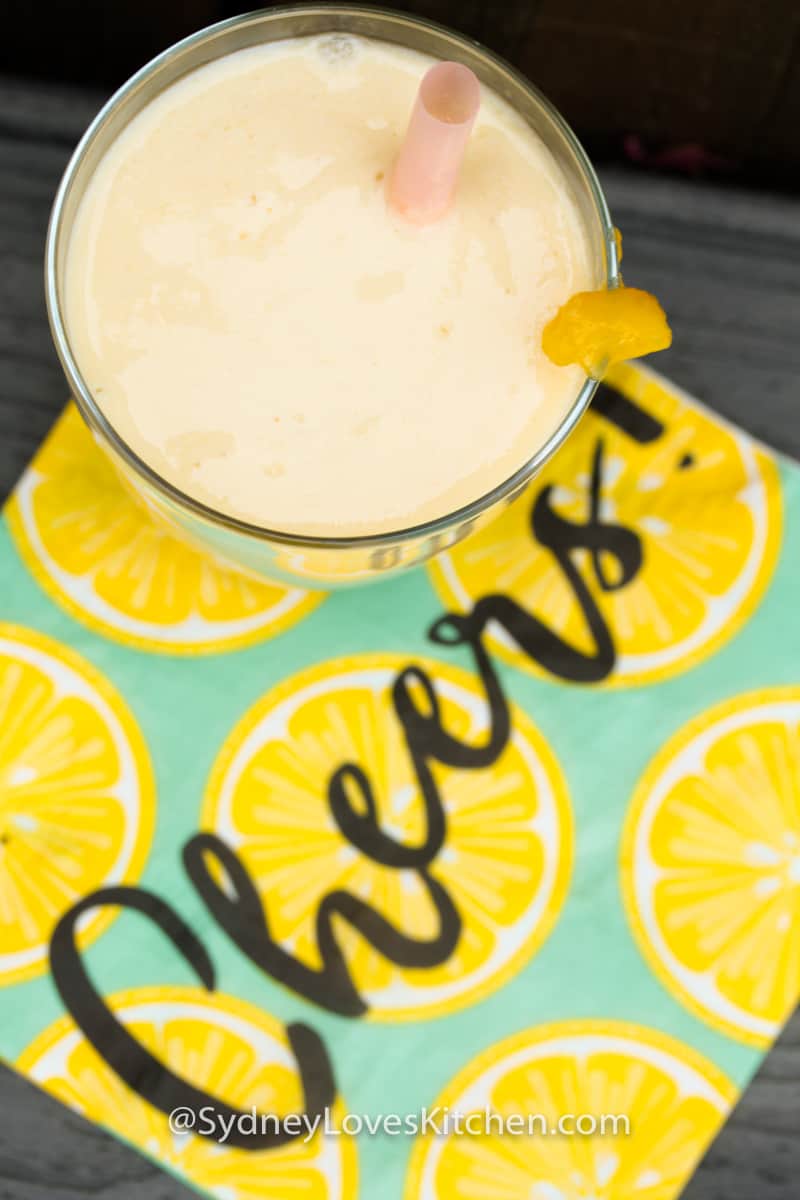 Overhead view of a glass of mango yogurt smoothie on top of a napkin that reads"cheers".